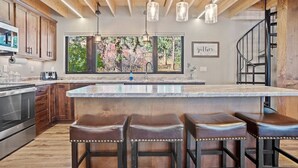 Una cocina moderna con armarios de madera, una isla grande con taburetes de bar, iluminación colgante y vistas a árboles a través de ventanas amplias.