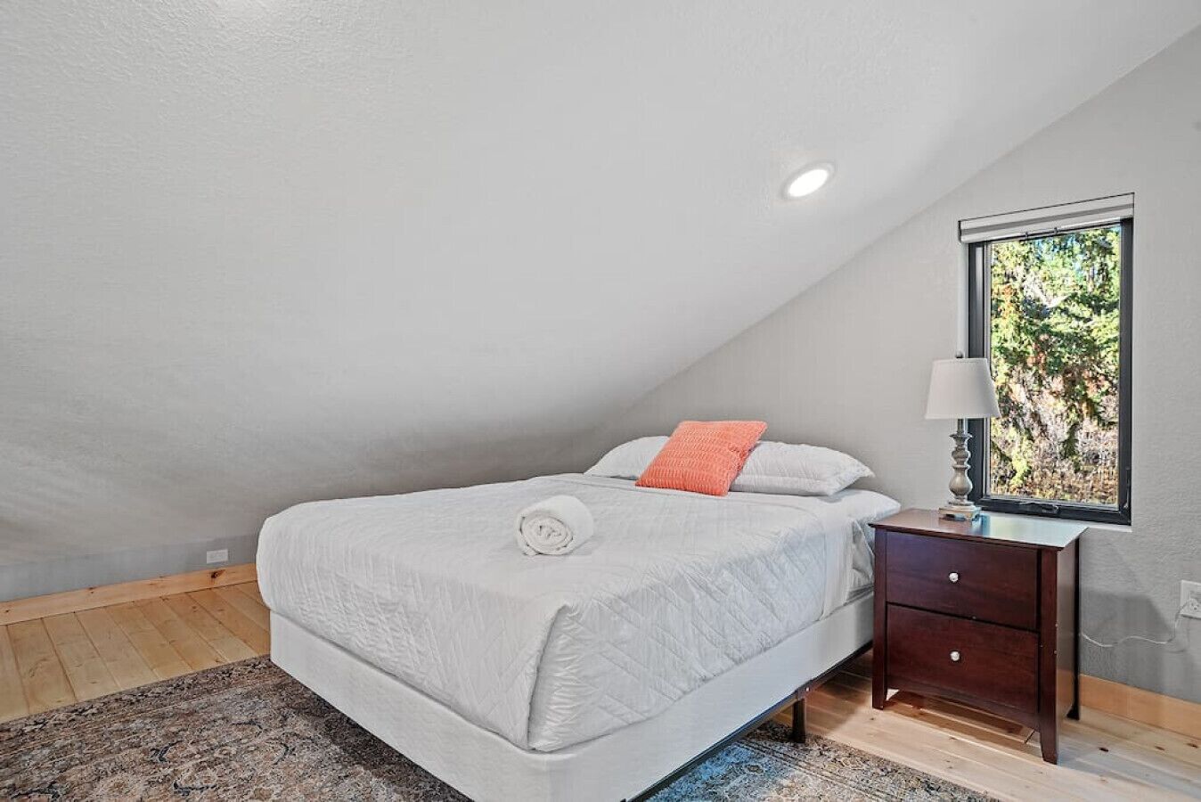 A bedroom with a bed, a nightstand, a lamp, and a window with a view of trees.