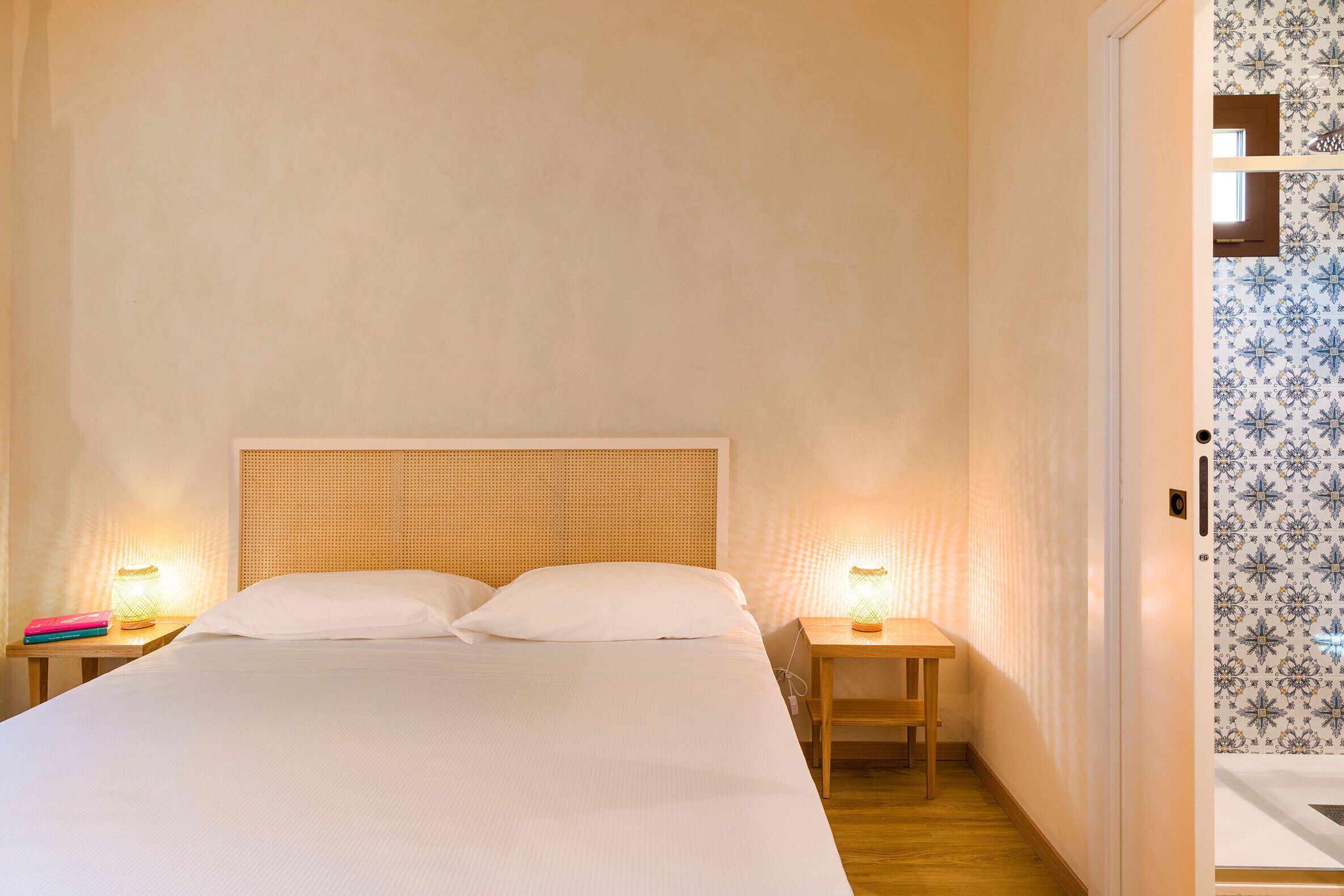 Ein ordentlich bezogenes Bett mit einem hölzernen Kopfteil, zwei Nachttische mit Lampen und einer gemusterten Wand rechts.