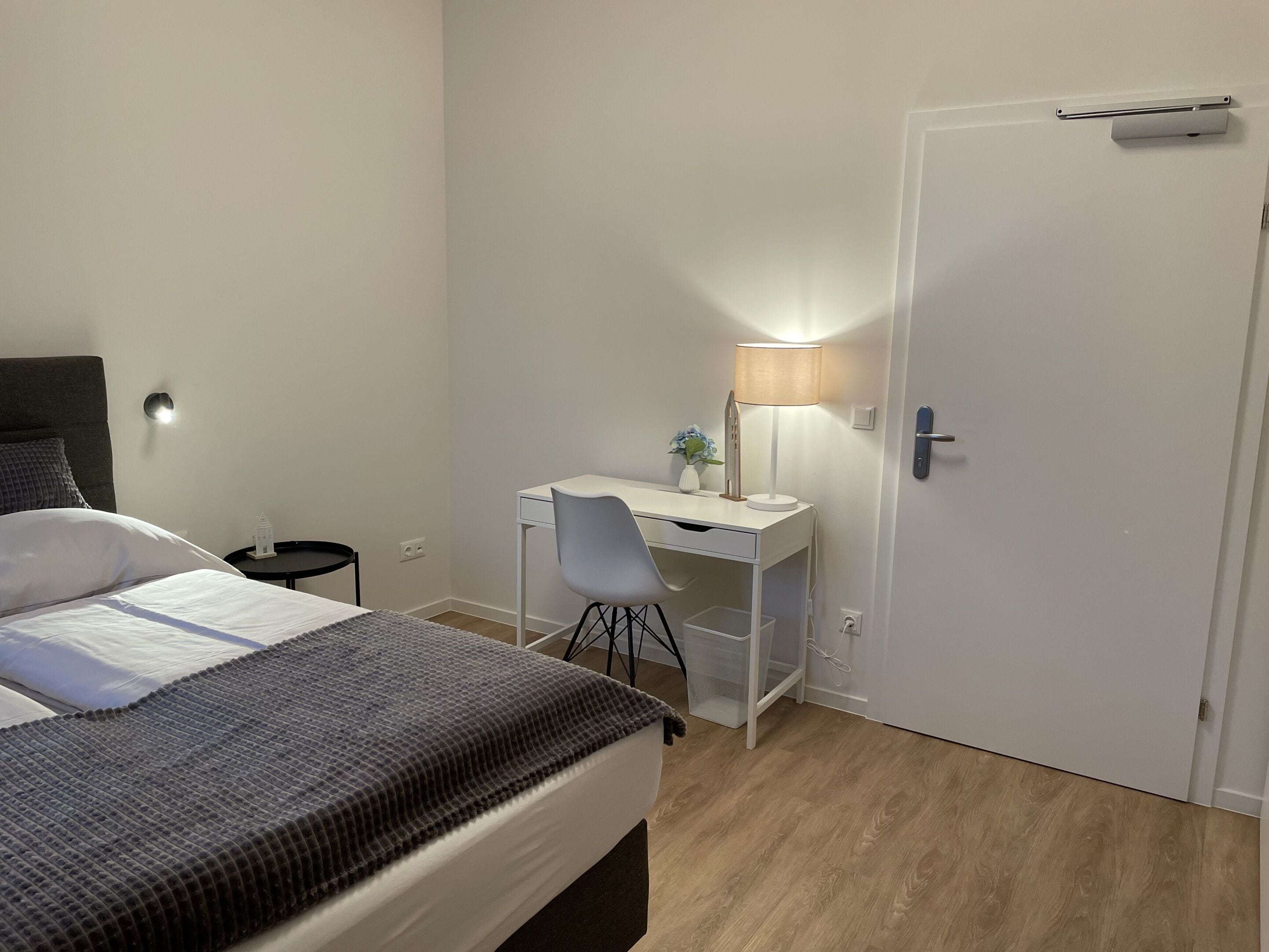 Ein Schlafzimmer mit Bett, Schreibtisch, Stuhl, Lampe und einer Tür.