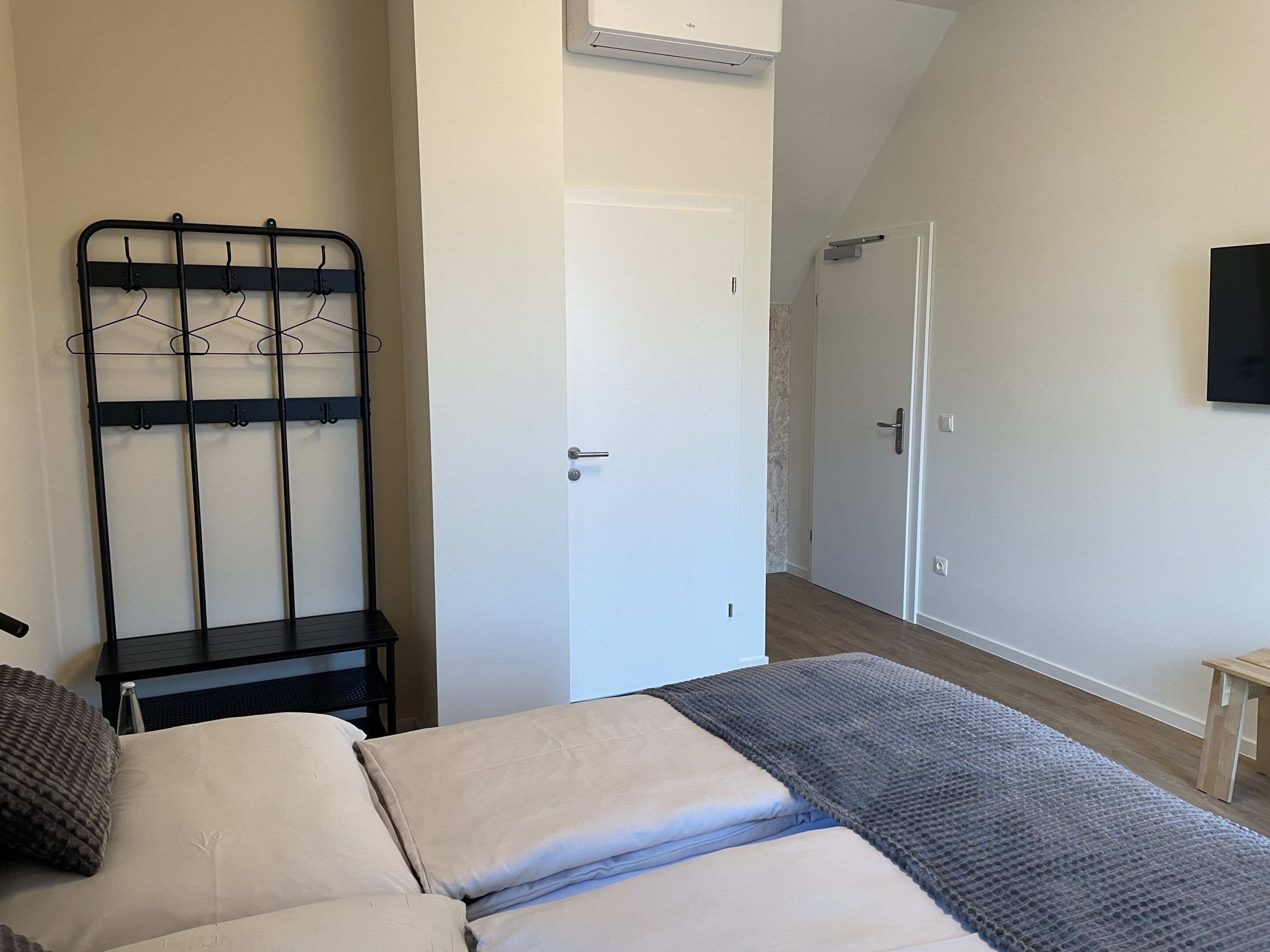 Quarto com cama, cabideiro de metal, porta e televisão na parede.
