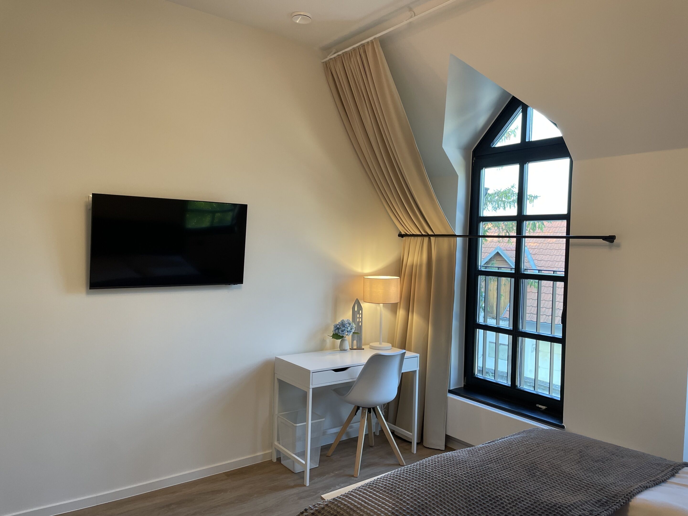 Un dormitorio con cama, escritorio, silla, televisor y ventana con cortinas.