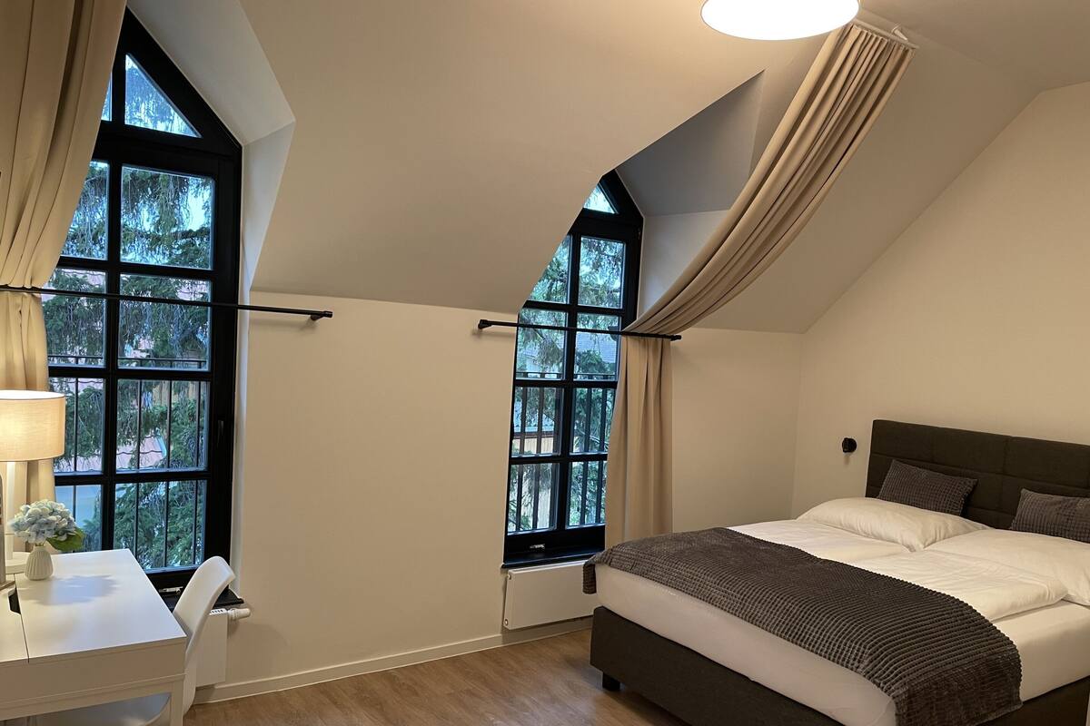 A bedroom with a bed, a desk, and two windows with curtains.