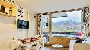 A cozy room with a dining table set for two, a television mounted on the wall, and a large window offering a mountain view. - Studio cabin with south balcony, 5 min to slopes, wifi included (Tignes)