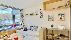 A cozy living room with a sofa, a dining table with food, a bookshelf, and a window with a city view. - Studio cabin with south balcony, 5 min to slopes, wifi included (Tignes)