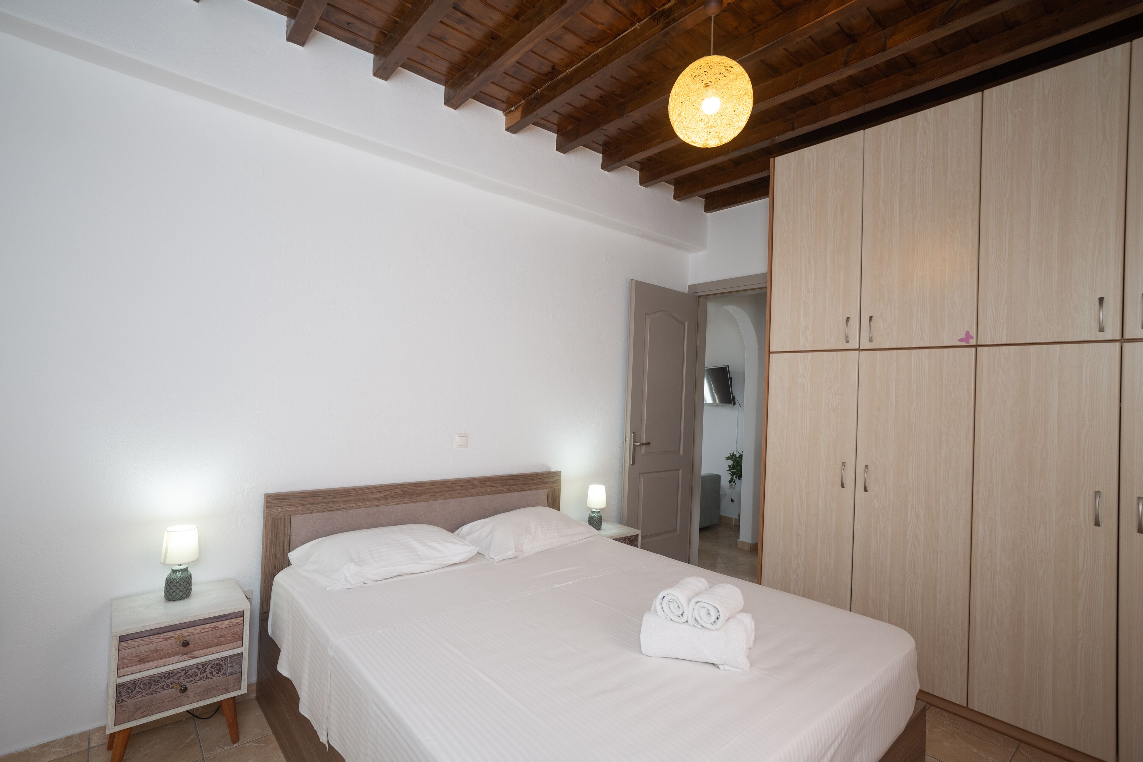 Un dormitorio con techo de madera, una cama con ropa blanca, dos mesitas de noche con lámparas y un armario.