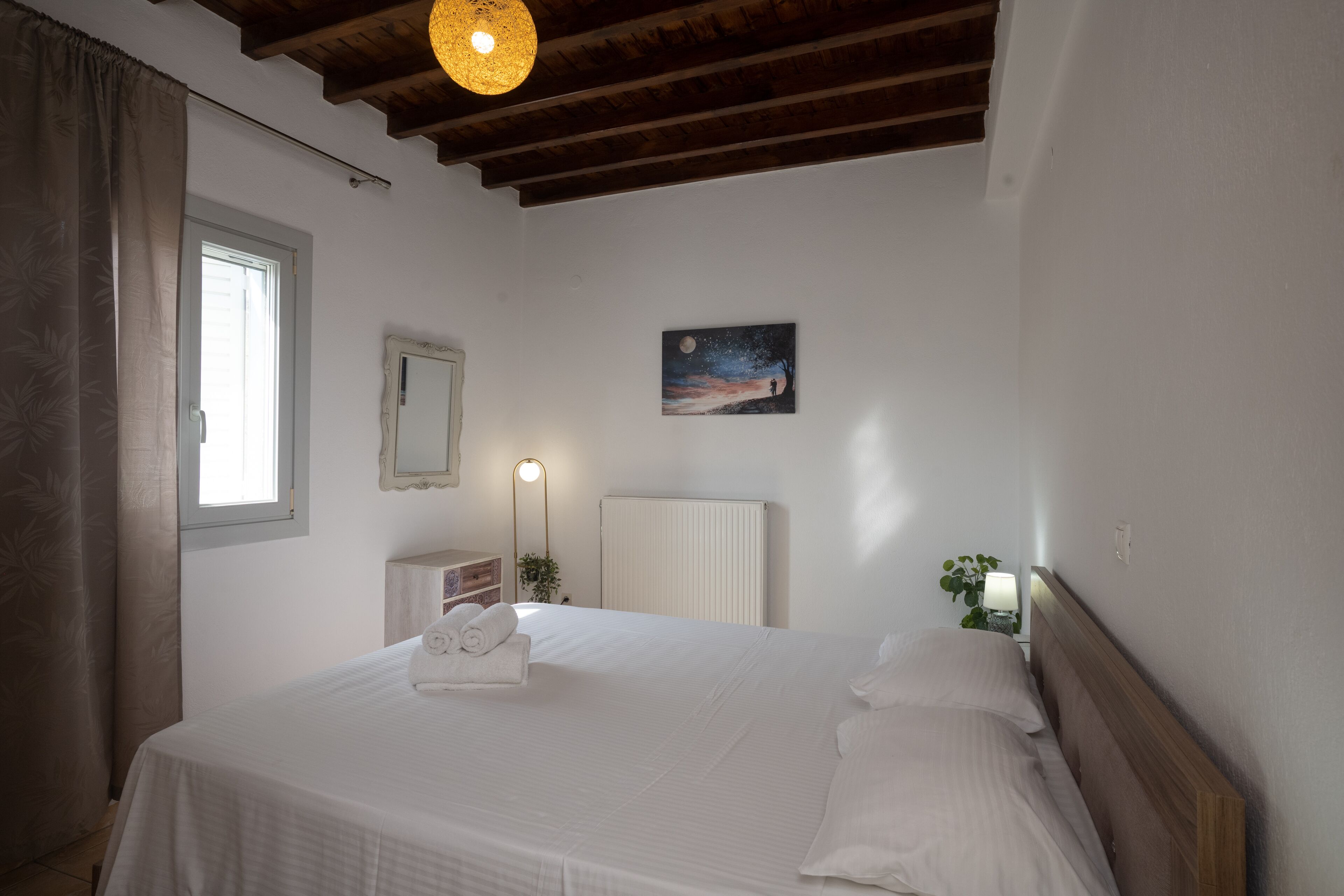 A neatly made bed with white linens and a rolled towel, a bedside lamp, a night sky painting on the wall, and a window with curtains.