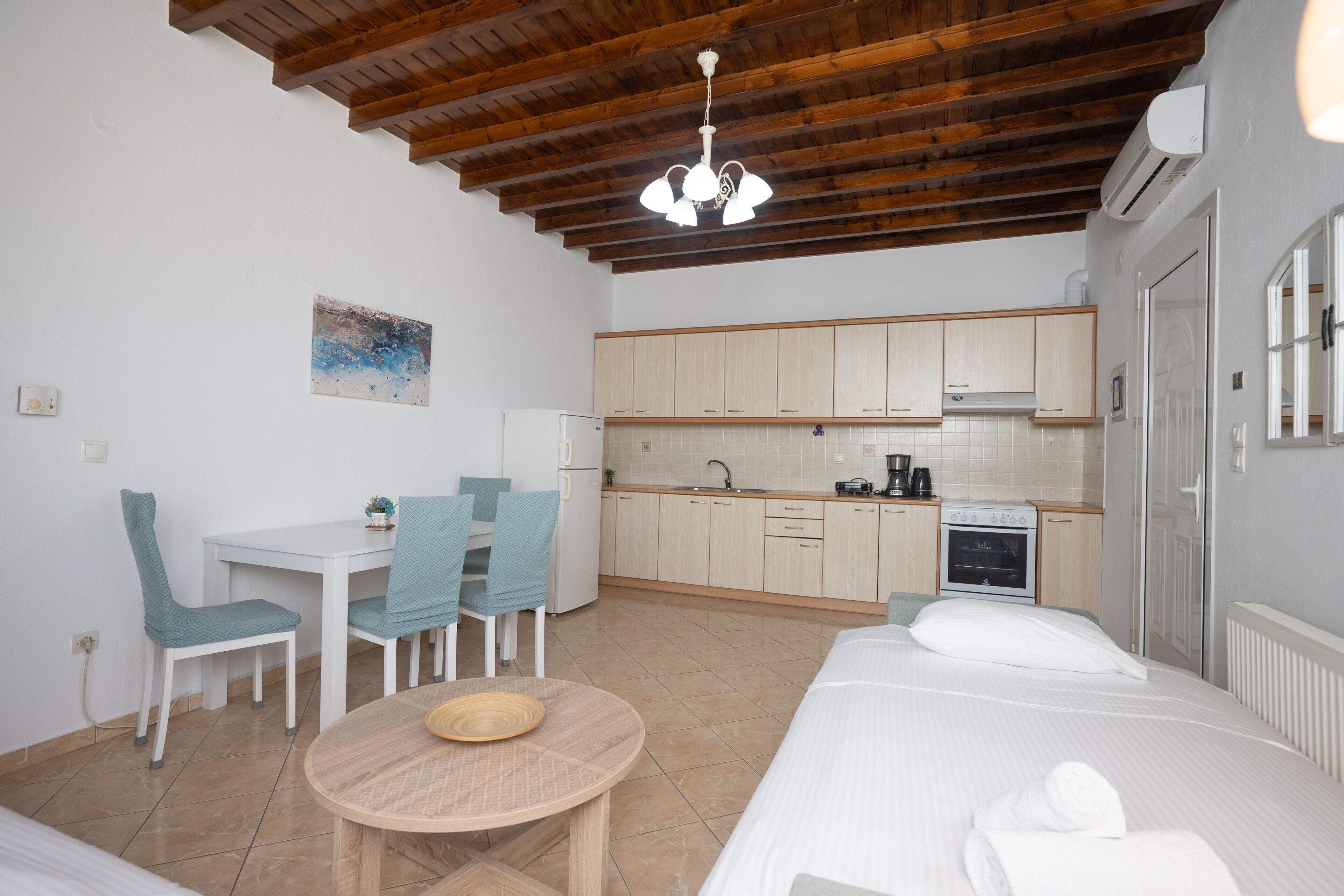Une chambre d’hôtel compacte avec un plafond en bois, une kitchenette, un coin repas avec une table et des chaises, et un lit avec des serviettes soigneusement pliées.