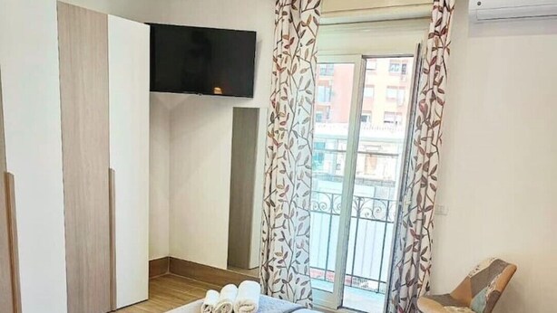 A hotel room with a bed, a chair, a TV, and a balcony with patterned curtains.