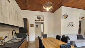 Une cuisine moderne avec un plan de travail en bois et un évier. On y trouve un coin repas avec une table en bois et des chaises noires. La pièce est dotée d’un plafond en bois et d’un luminaire fixé au mur.