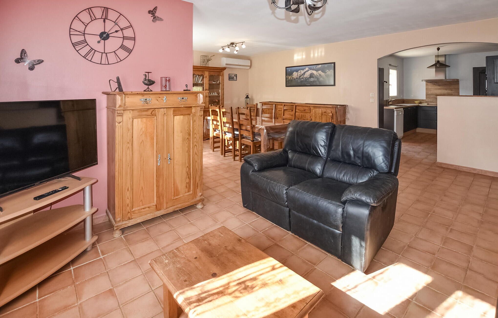A living room with a TV, wooden cabinet, leather sofa, and a dining area with chairs and a table.