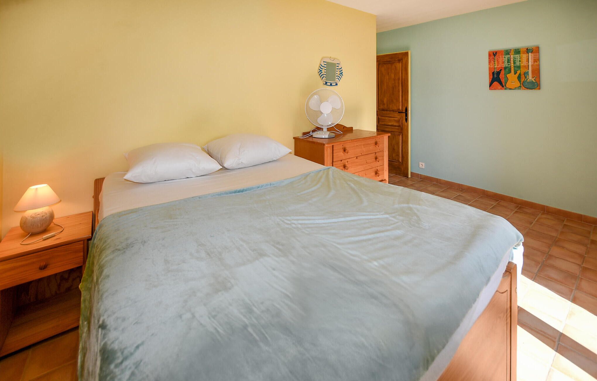 Un dormitorio con una cama, una mesita de noche, un aparador, un ventilador y un cuadro con temática de guitarra en la pared.