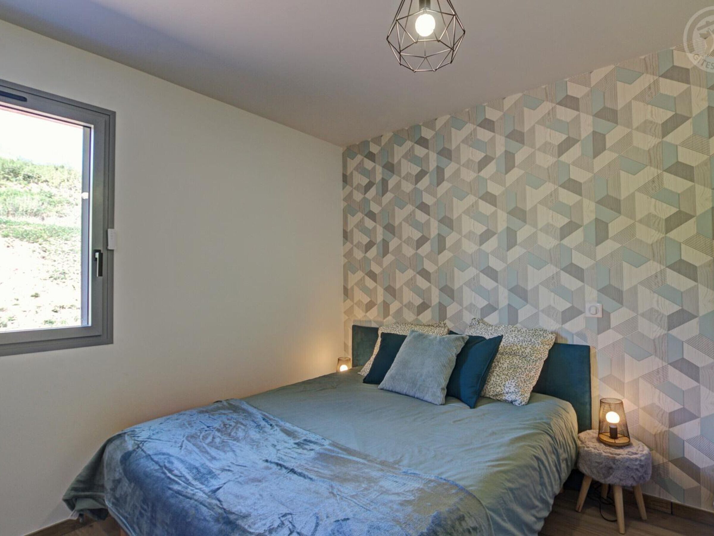 A bedroom with a bed, a geometric patterned wall, a small bedside table with a lamp, and a window with a view of greenery.