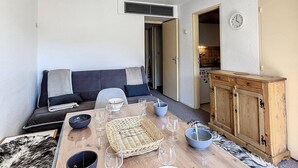 Un salon chaleureux avec un canapé, une table à manger dressée avec des verres et des bols, un buffet en bois et une cuisine visible à travers une porte ouverte.