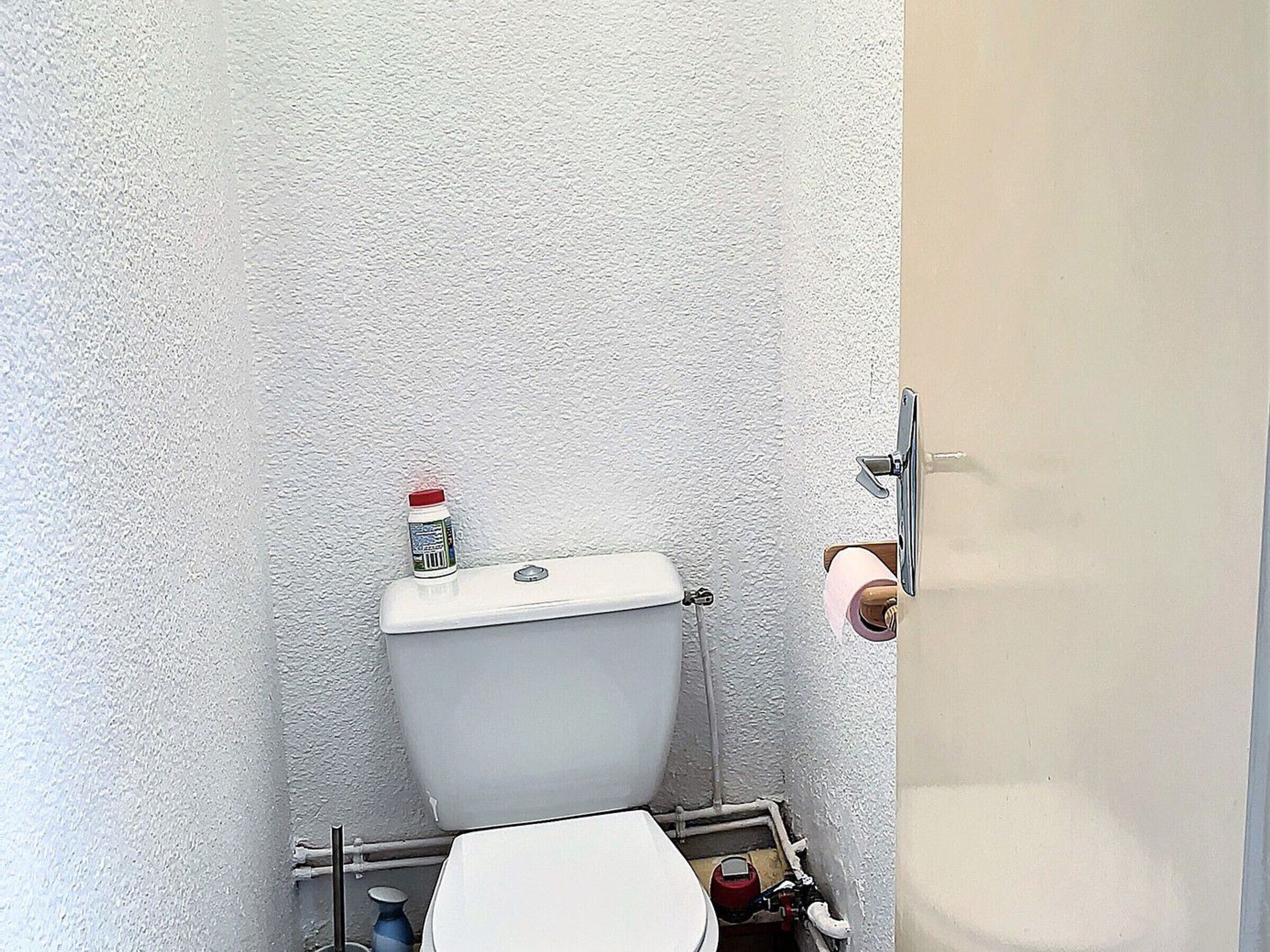 A small, compact bathroom with a white toilet, a small bottle of cleaning product on the tank, and a toilet paper holder on the wall.