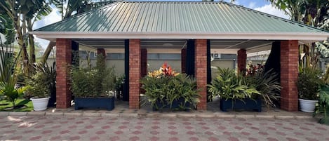 A covered outdoor area with a tiled floor, surrounded by potted plants and palm trees.