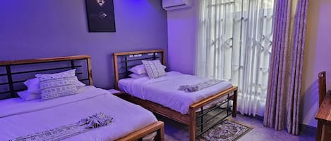 A room with two wooden beds, a wall-mounted air conditioner, and a window with sheer curtains.