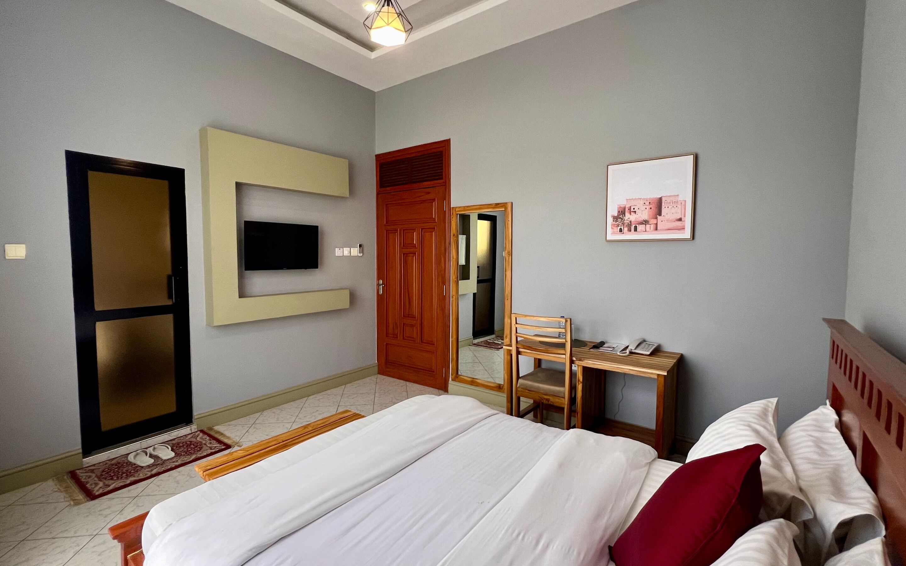 A hotel room with a bed, a desk with a chair, a television mounted on the wall, and a door leading to another room.