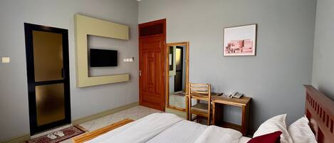 A hotel room with a bed, a desk with a chair, a television mounted on the wall, and a door leading to another room.