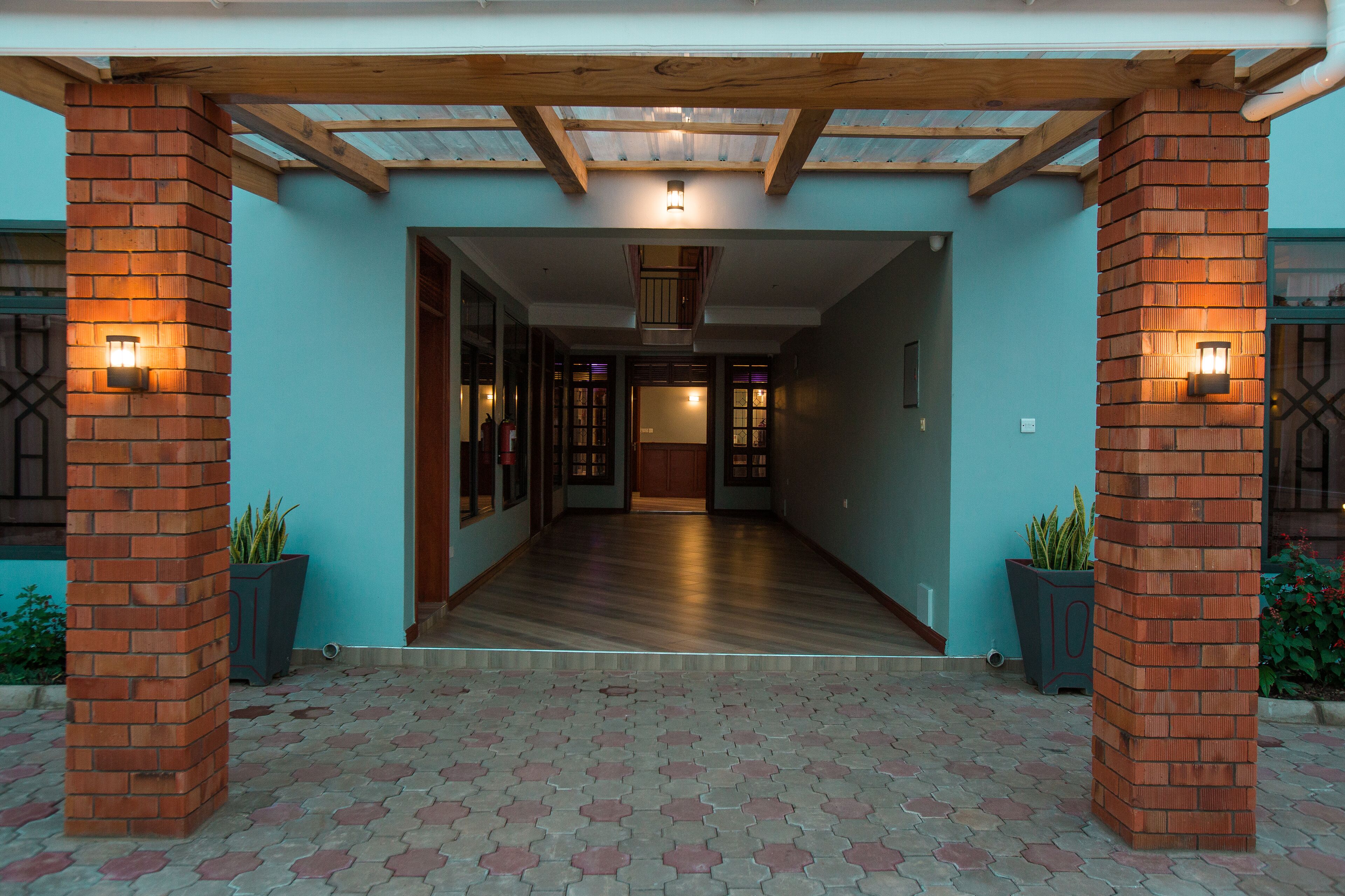 Una entrada techada con techo de madera y pilares de ladrillo, que conduce a un pasillo con piso de madera y vistas al interior del edificio.