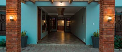 Una entrada techada con techo de madera y pilares de ladrillo, que conduce a un pasillo con piso de madera y vistas al interior del edificio.