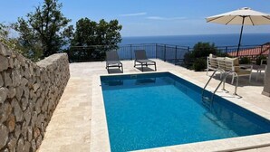 A rectangular swimming pool with clear blue water, surrounded by stone walls and a stone-paved deck. There are lounge chairs and a table with an umbrella on the deck. - Family Vacation Villa Rezevici (Petrovac)