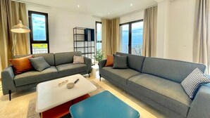 A modern living room with two grey sofas, a white coffee table, and a blue ottoman. There is a bookshelf and a large window with a view. - Family Vacation Villa Rezevici (Petrovac)