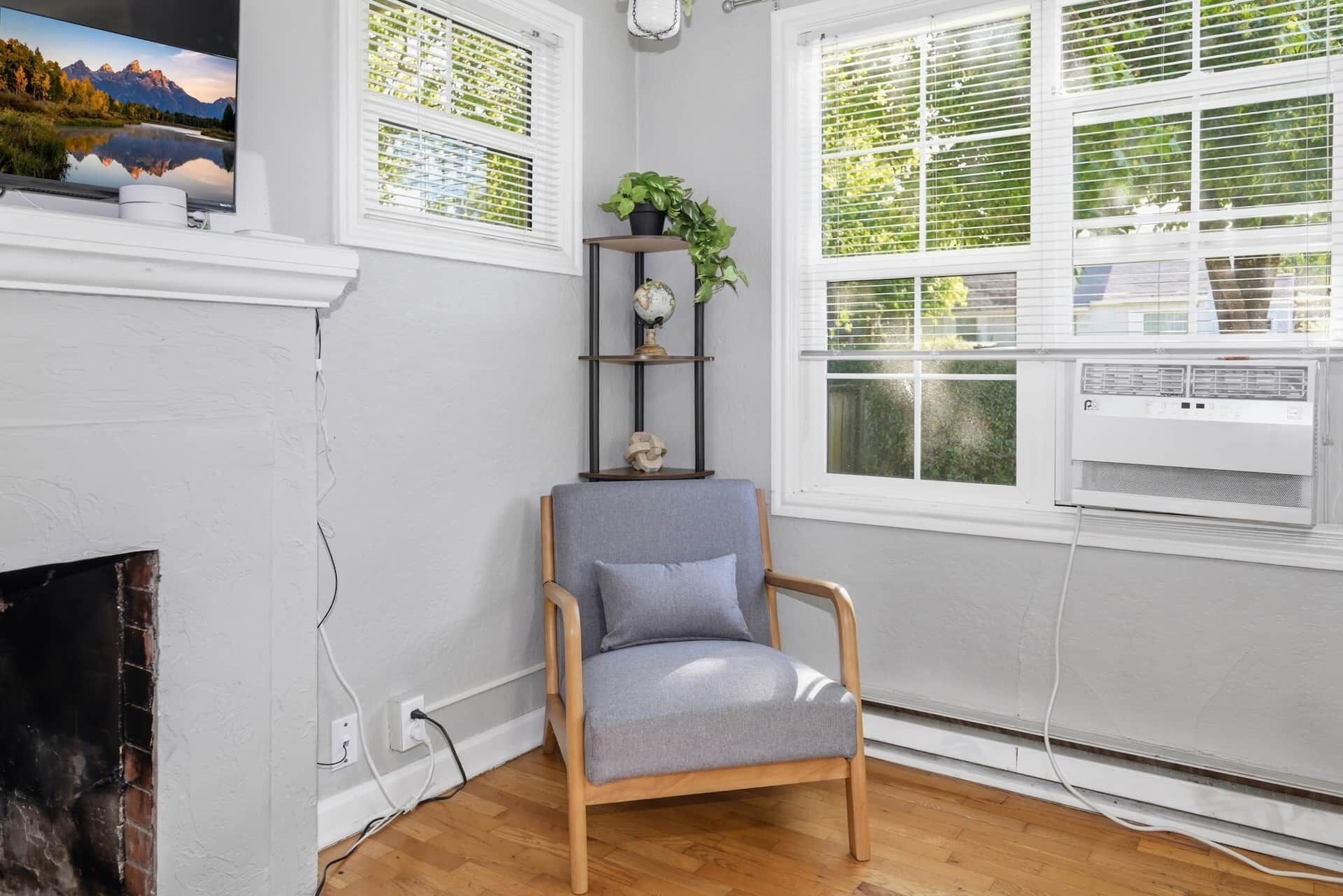 Een kamer met een houten vloer, een grijze fauteuil, een open haard en een groot raam met zonwering.