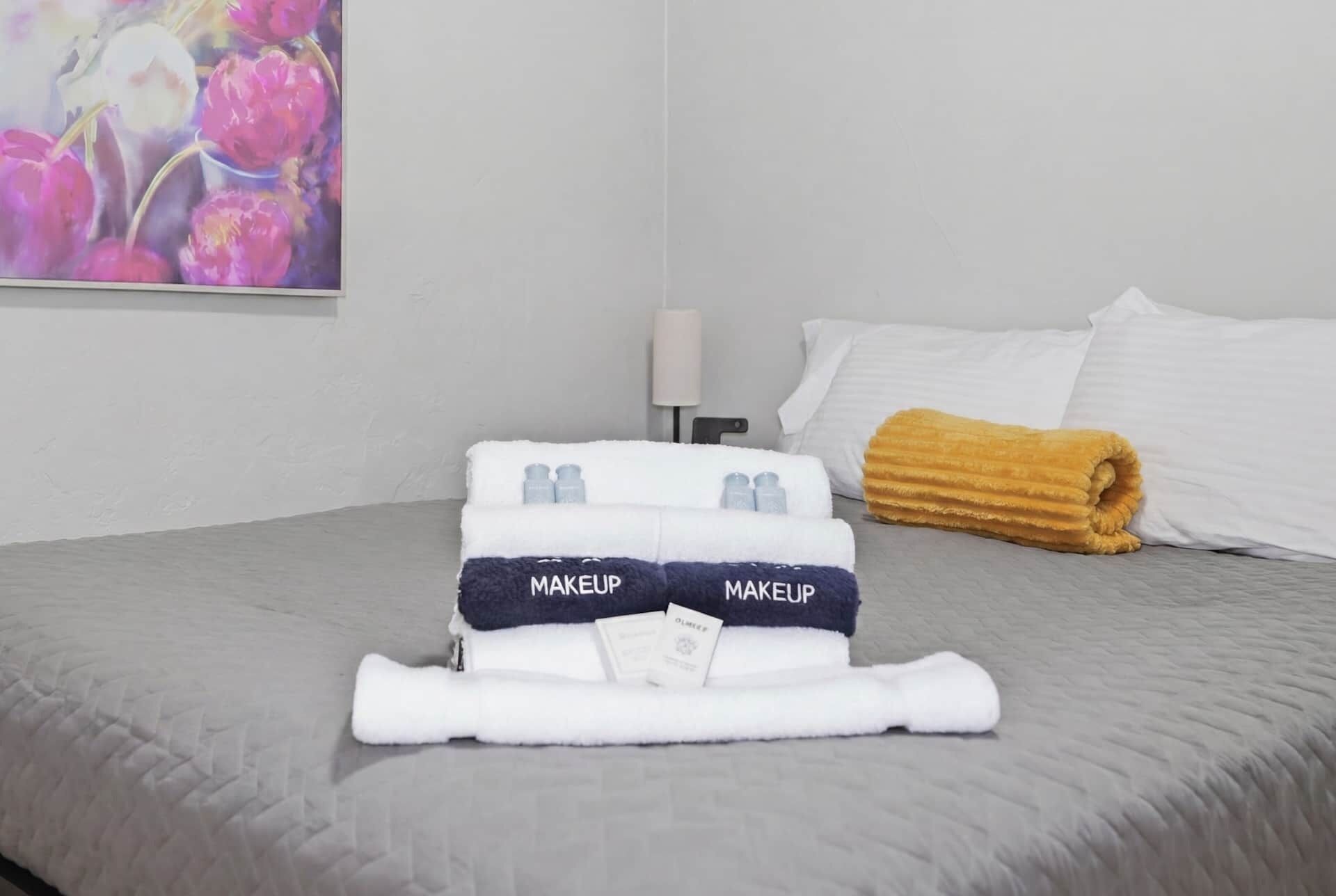 A neatly made bed with a folded towel labeled 'MAKEUP' and a rolled towel on the side.