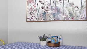 A room with a checkered tablecloth, a painting of flowers on the wall, and a small table with snacks and water bottles. - Charming 1930s Style Rustic Home (Eugene)