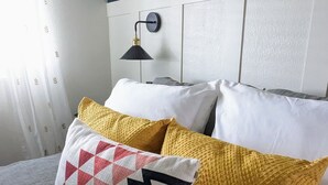 A bedroom with a bed, pillows, a nightstand, a wall-mounted lamp, and a shelf with plants. - Quiet and Unique 2BR on Winery Row - The Elgin Project (Elgin)