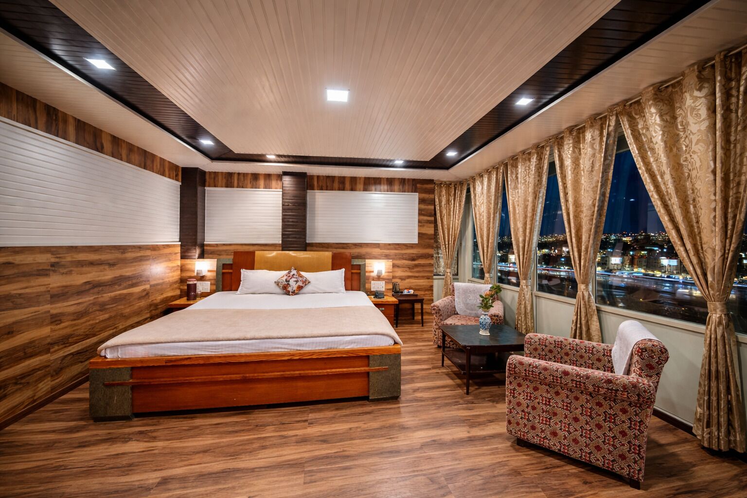 Una habitación de hotel con una cama grande, una mesita, una silla y vistas de la ciudad de noche.