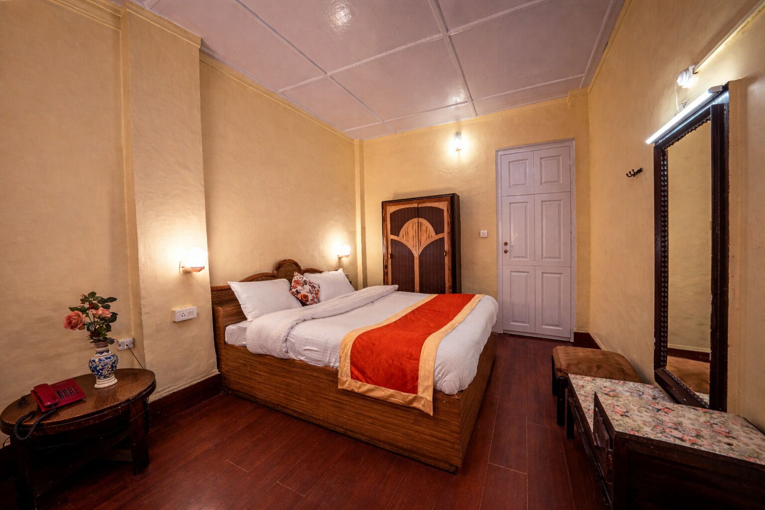 A bedroom with a wooden bed, bedside lamps, a nightstand with a vase, a wardrobe, and a mirror.