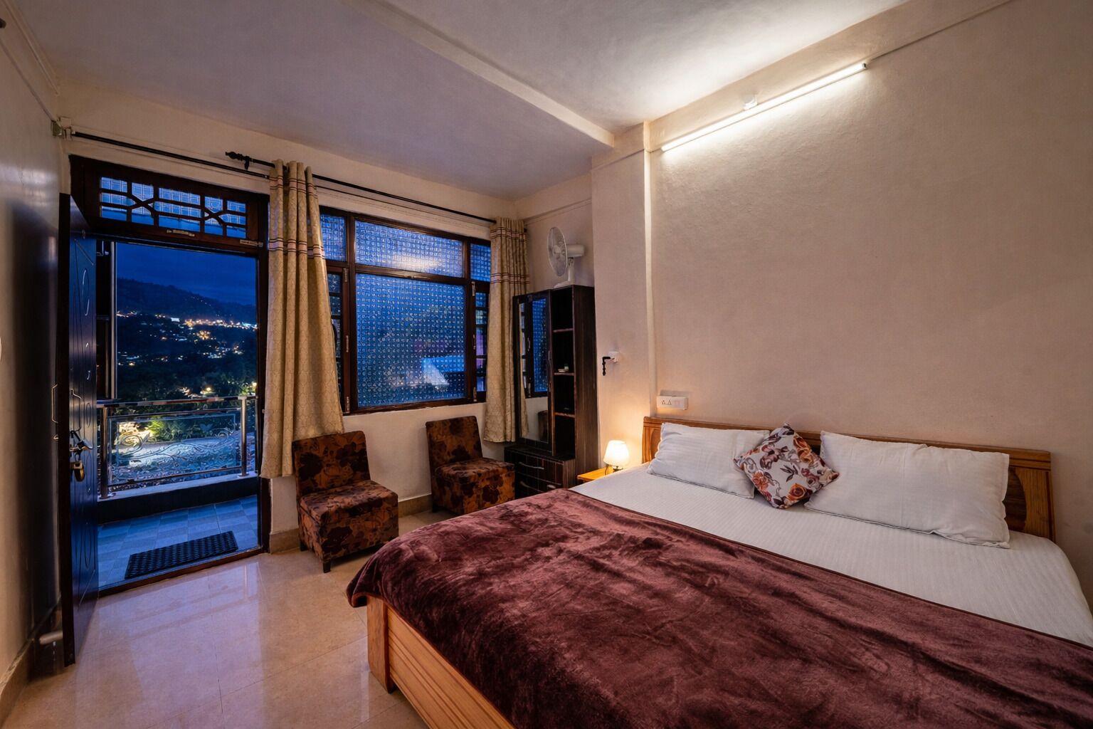 Un dormitorio con una cama grande, una mesita de noche, una lámpara, una ventana con cortinas, vista a un paisaje urbano y una pequeña zona de estar.