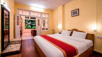 Hotelkamer met een groot bed, een houten hoofdeinde, een rode spreideken, een klein tafeltje met stoelen en uitzicht op groen door de open deur.