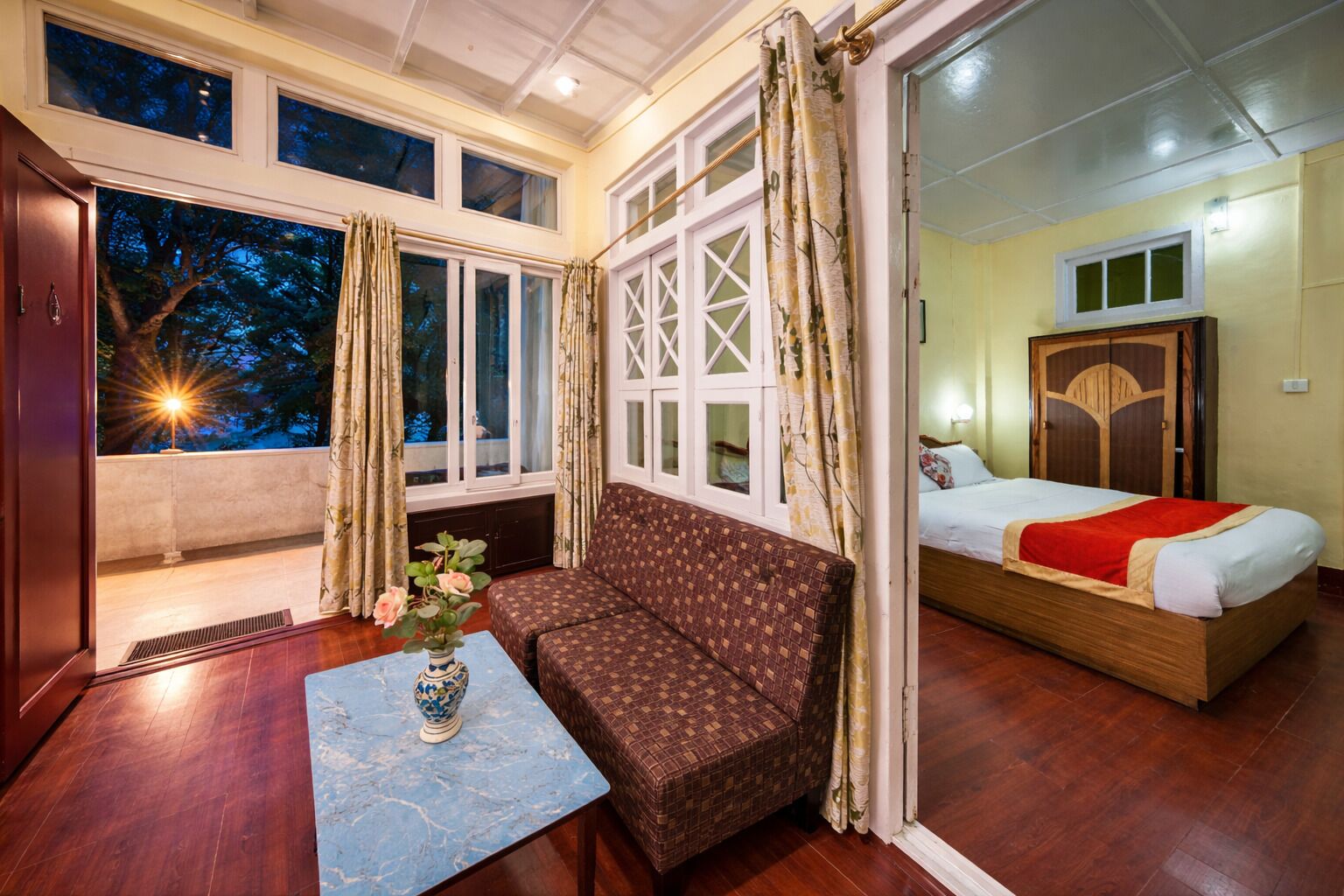 A room with a sofa, a bed, a window with curtains, and a vase with flowers on a table.