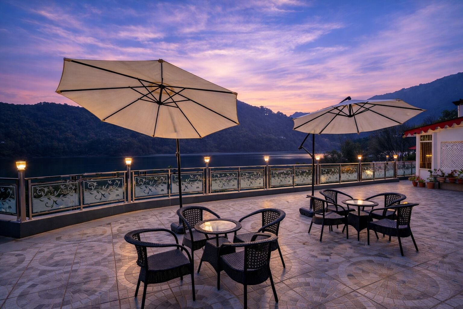 An outdoor terrace with tables and chairs, large umbrellas, and a view of a lake at dusk.