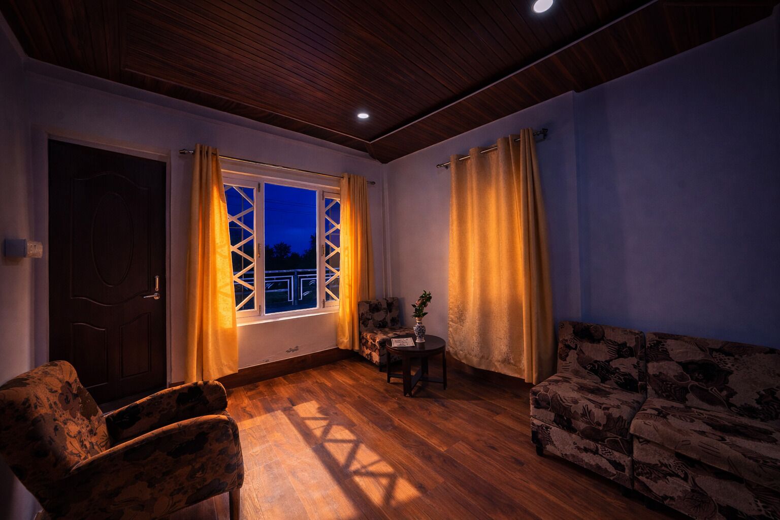Una habitación con techo de madera, ventana con cortinas, sofá, una silla y una pequeña mesa con una planta.