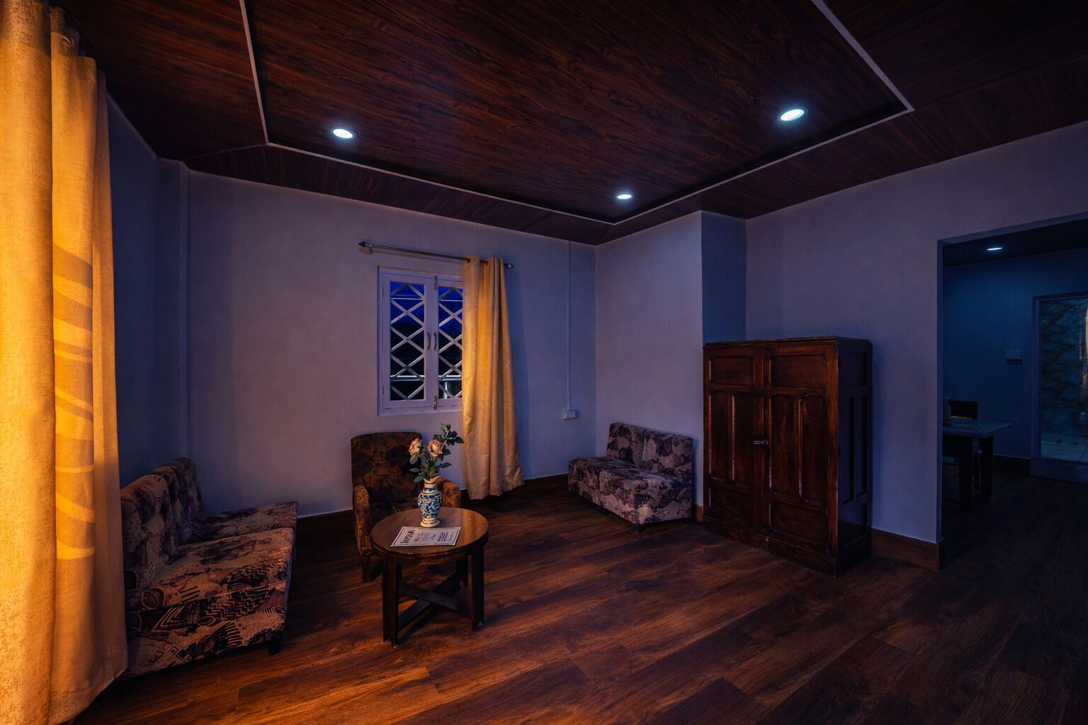 A room with a wooden ceiling, a window with curtains, two patterned armchairs, a small table with a vase, and a wooden cabinet.