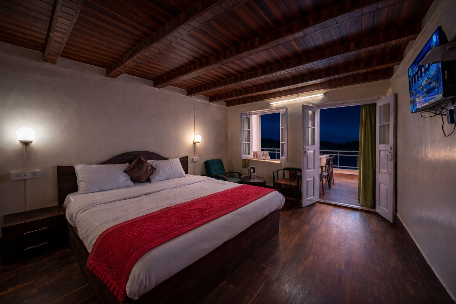 A bedroom with a wooden ceiling, a bed with white bedding and a red throw, bedside lamps, a TV on the wall, and a balcony with chairs.