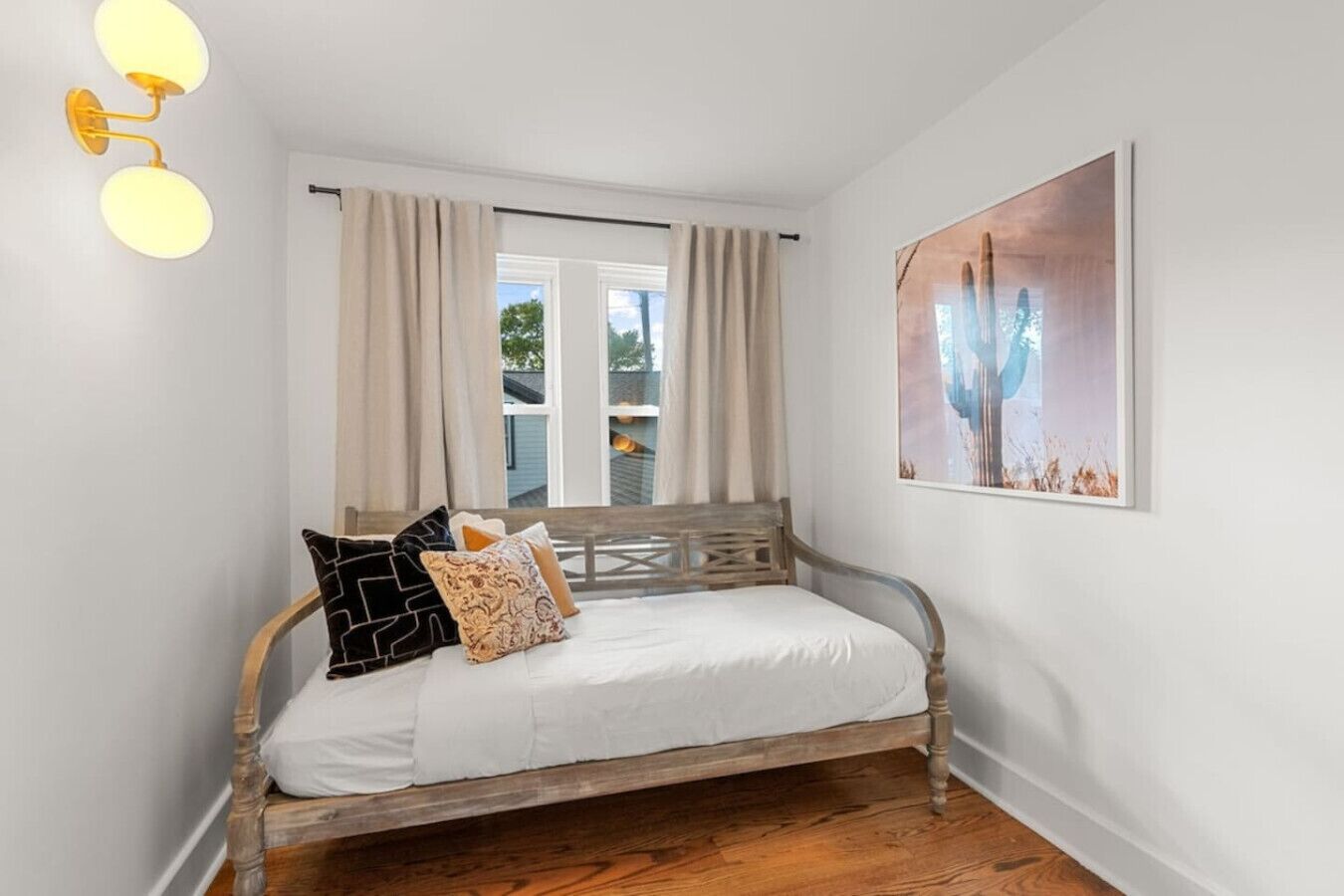 A bedroom with a wooden bed, beige curtains, a window with a view, and a painting on the wall.