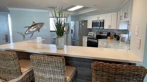 A modern kitchen with white cabinets, stainless steel appliances, and a central island with wicker chairs. - Gulf Front Condo with Stunning Views! (Pensacola)