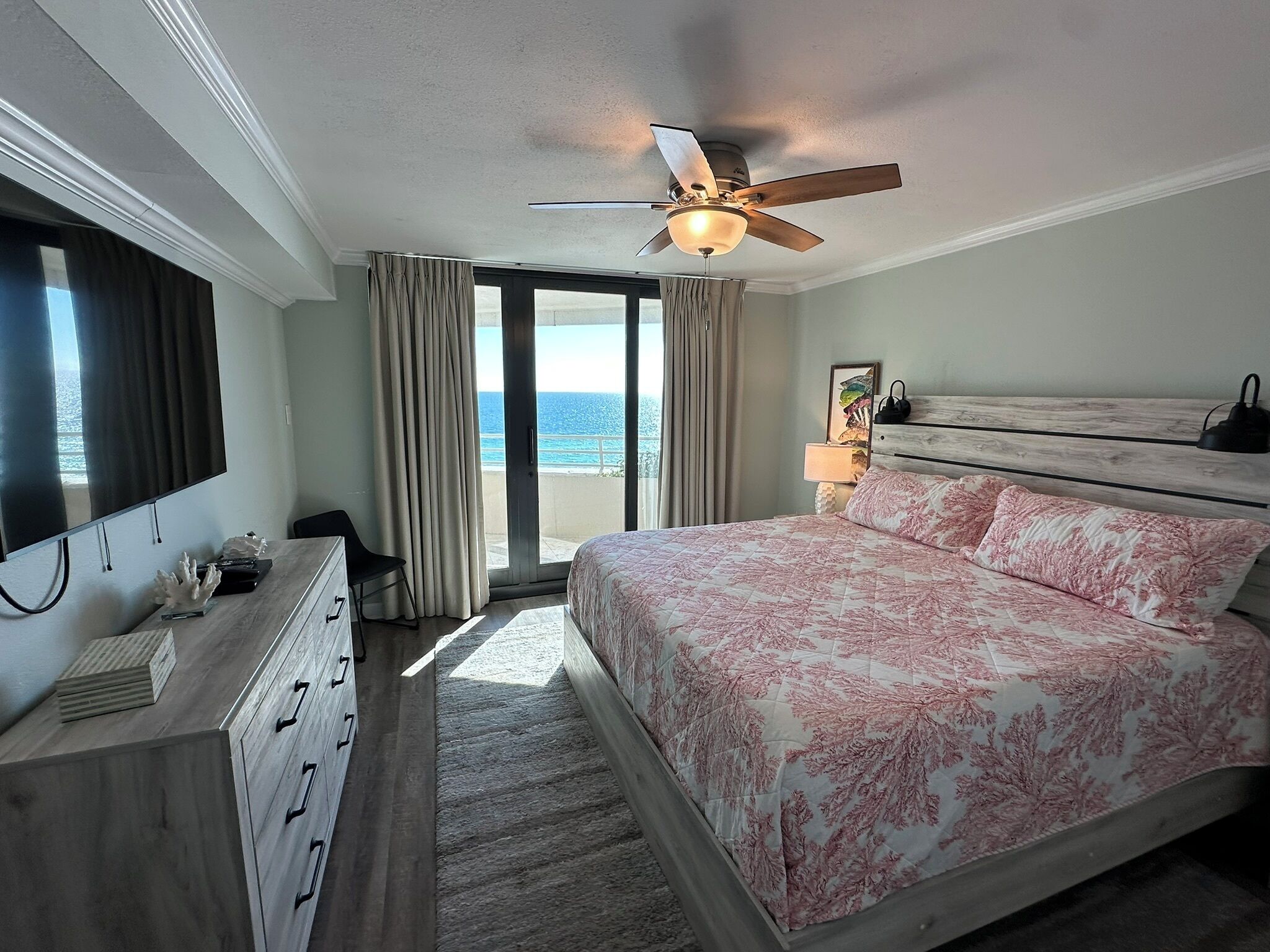 Un dormitorio con cama, ventilador de techo, televisor, cómoda y vista al océano.