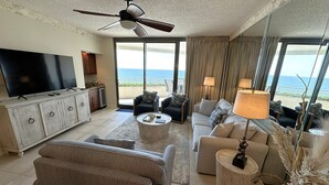 A spacious living room with a large flat-screen TV, comfortable seating, a ceiling fan, and a view of the ocean through large windows.