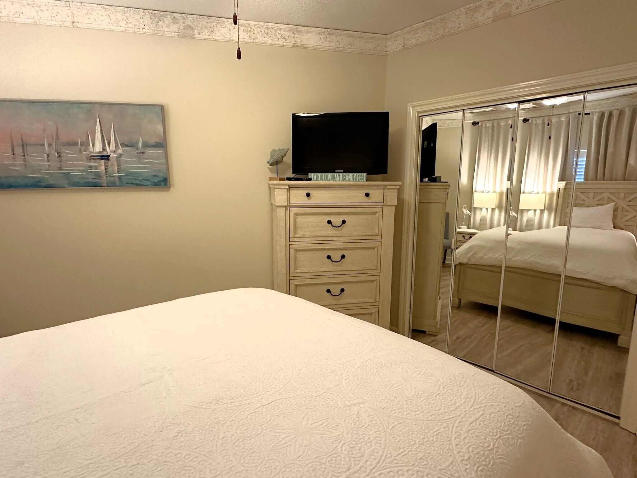 A hotel room with a bed, a dresser with drawers, a television on top, a painting of sailboats, and a mirror with curtains.