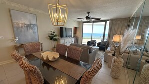 A modern living room with a dining area, large windows, a ceiling fan, and a view of the ocean.