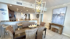 A dining area with wicker chairs and a central table, a kitchen with a marble countertop, and a large abstract painting on the wall.