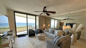 A spacious living room with a large sofa, armchairs, and a coffee table. There is a ceiling fan, a TV, and a view of the ocean through large windows.