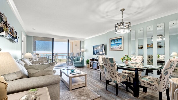 A spacious living room with a sofa, coffee table, and patterned chairs. A dining table with chairs is in the center. Large windows offer a view of the outdoors. - FAMILY FAVORITE!! NEW UPGRADES!  Booking for Summer NOW! (Myrtle Beach)