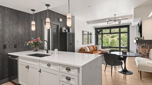 Una cocina moderna con una isla central, paredes oscuras e iluminación colgante. Hay una zona de comedor con una mesa redonda y sillas, y un sofá al fondo.